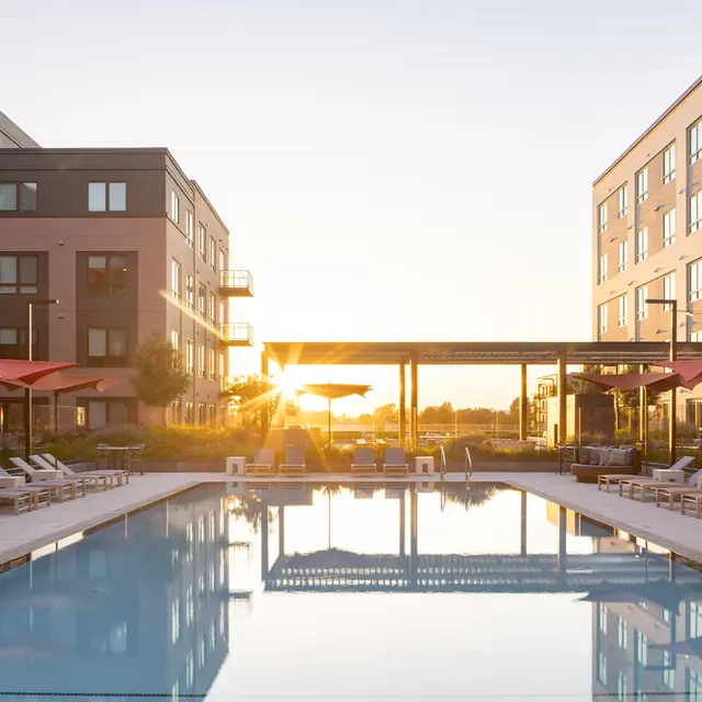 A serene pool area at sunset, surrounded by modern apartment buildings. The pool reflects the colorful sky as the sun sets in the background, with lounge chairs and umbrellas lining the poolside.