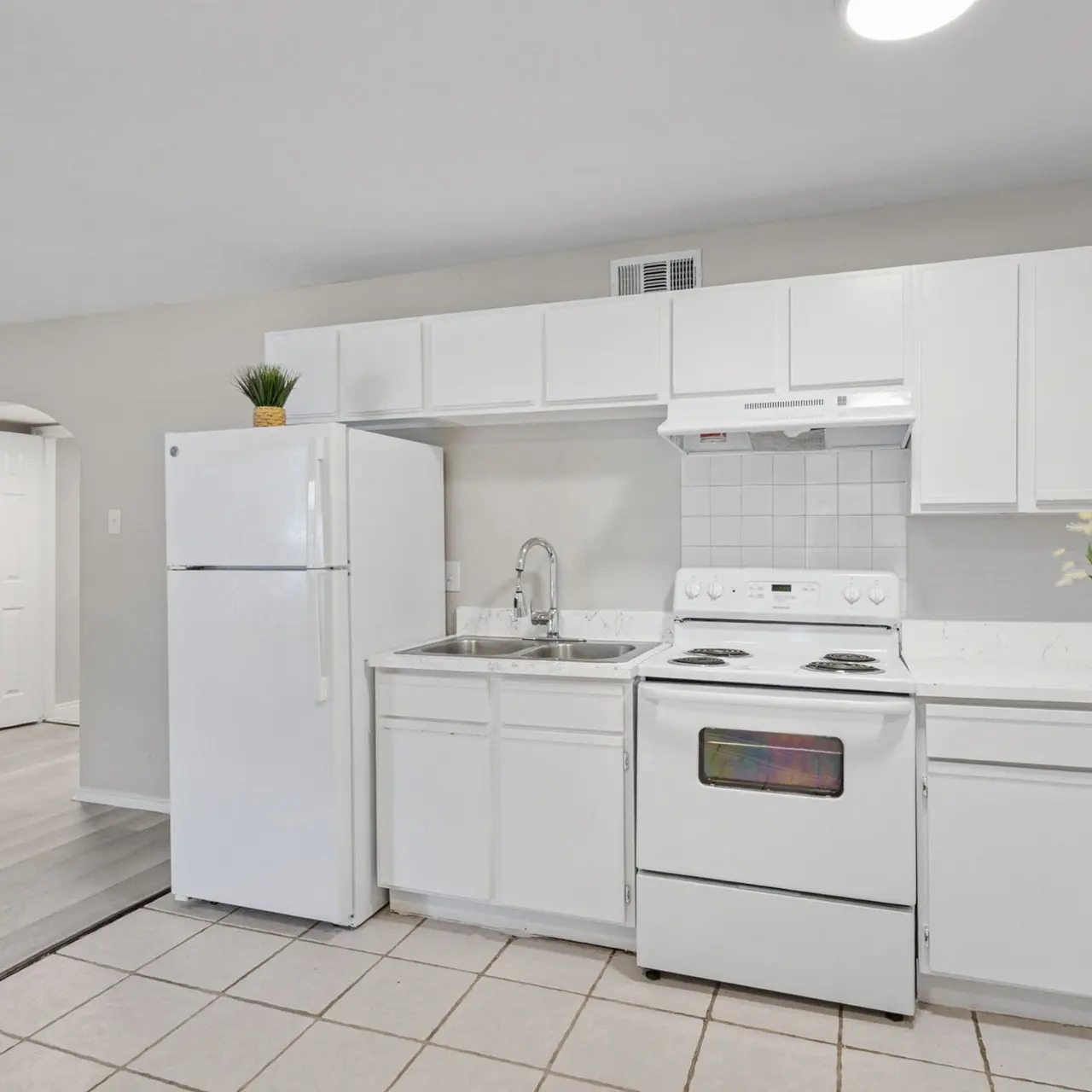 An expansive kitchen with white cabinetry, a white refrigerator, and a white stove. The space features tiled flooring and a bright, natural lighting. A vase of flowers sits on the counter next to the sink. A doorway in the background leads to another room.