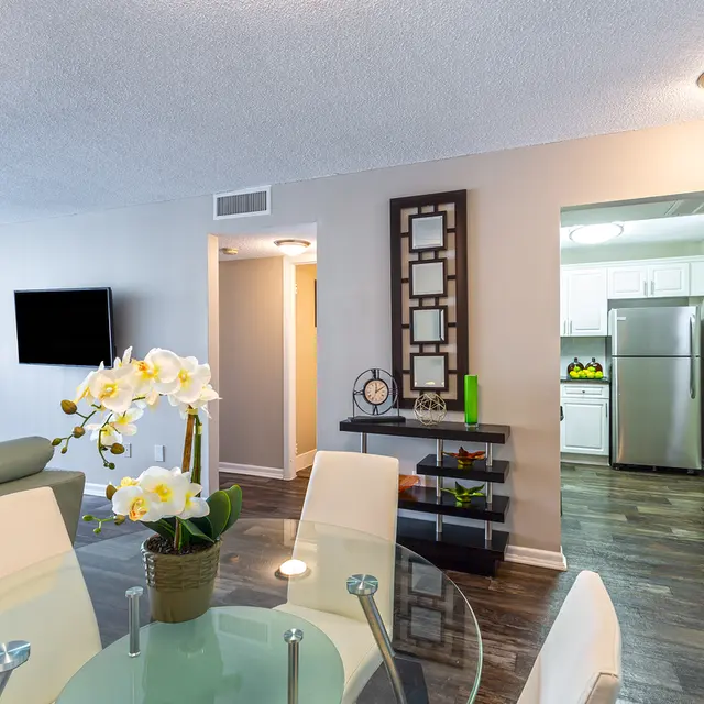 A modern apartment interior featuring a glass dining table with white chairs, a decorative orchid plant, and a living area visible in the background with a flat-screen TV and access to a kitchen.