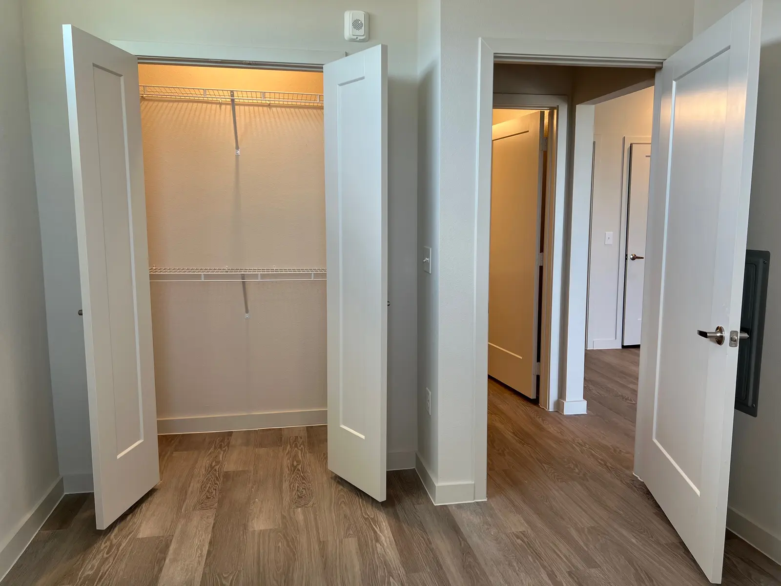 A room featuring two open closet doors on the left, revealing a wire shelf inside. To the right, a hallway can be seen with another door partially visible. The flooring is a light wood-like material, and the walls are painted a light color.