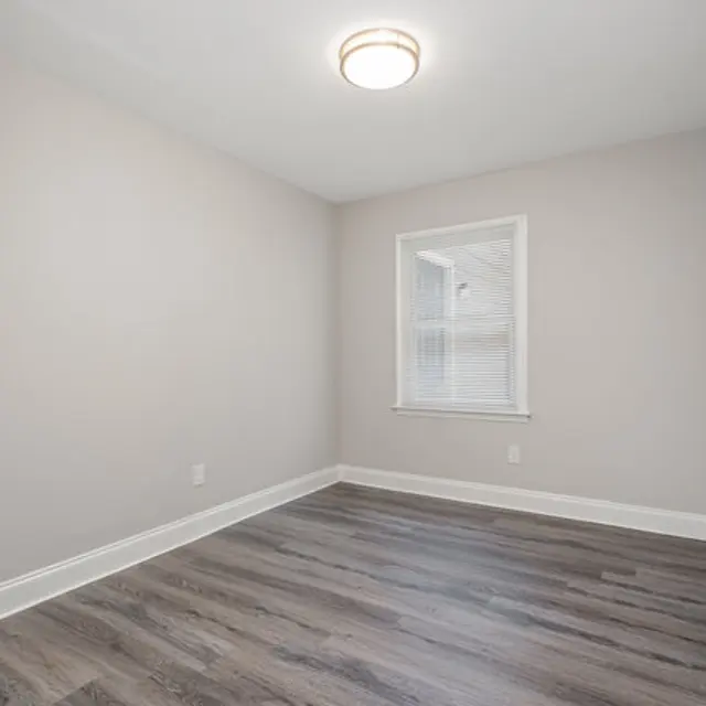 A small, empty room with light gray walls and laminate flooring. A single window is visible, providing natural light. The ceiling features a circular light fixture.