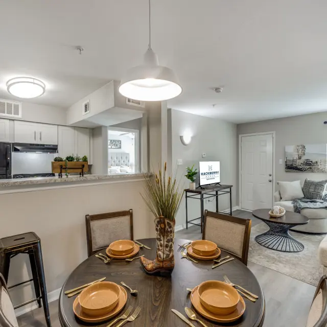 A modern apartment dining area with a round table set for four, featuring plates and utensils. In the background, there is a kitchen with black appliances and a cozy living room with a sofa and coffee table. Bright natural light comes through the window, enhancing the elegant decor.