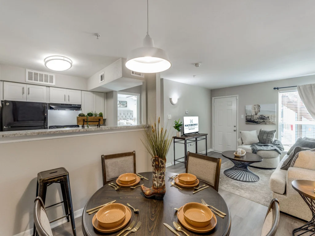 A modern apartment dining area with a round table set for four, featuring plates and utensils. In the background, there is a kitchen with black appliances and a cozy living room with a sofa and coffee table. Bright natural light comes through the window, enhancing the elegant decor.