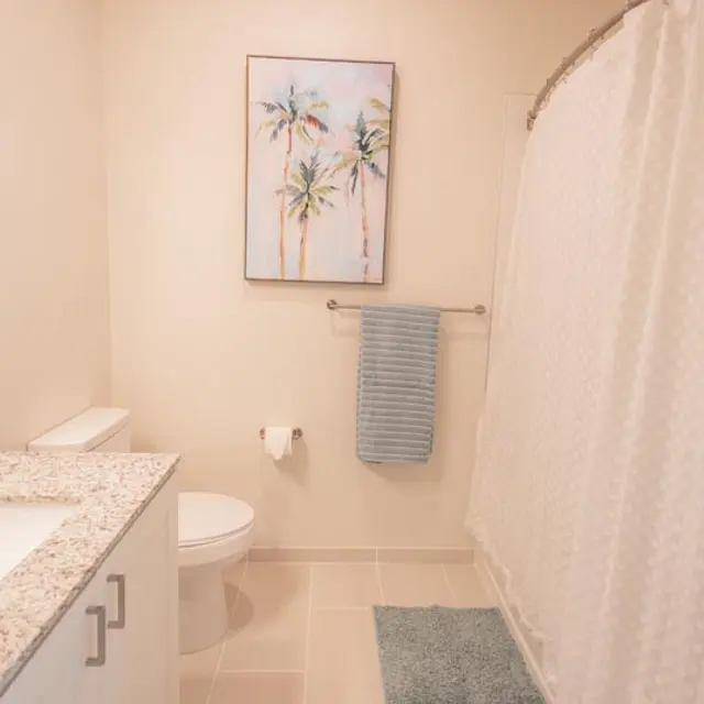 A modern bathroom featuring a light-colored tiled floor, a large mirror above a granite countertop with white cabinetry, a shower curtain, and a small artwork of palm trees on the wall.