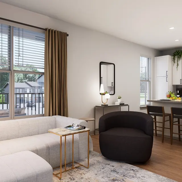 A modern living room with a light-colored sectional sofa, a dark armchair, a coffee table, and a lamp. The room features large windows with brown curtains and wooden flooring, leading to an open kitchen with white cabinets and a bar with stools.