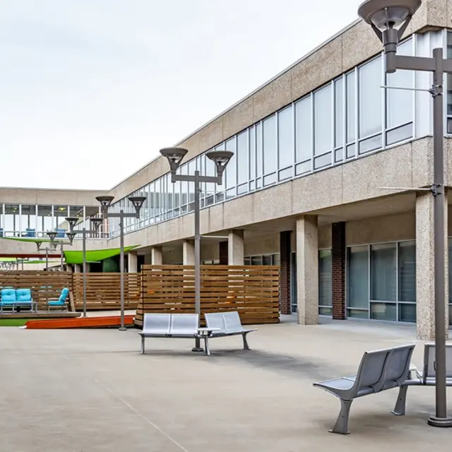 A modern outdoor courtyard featuring concrete architecture with benches, lamps, and a grass area. In the background, colorful furniture can be seen, along with wooden fencing.