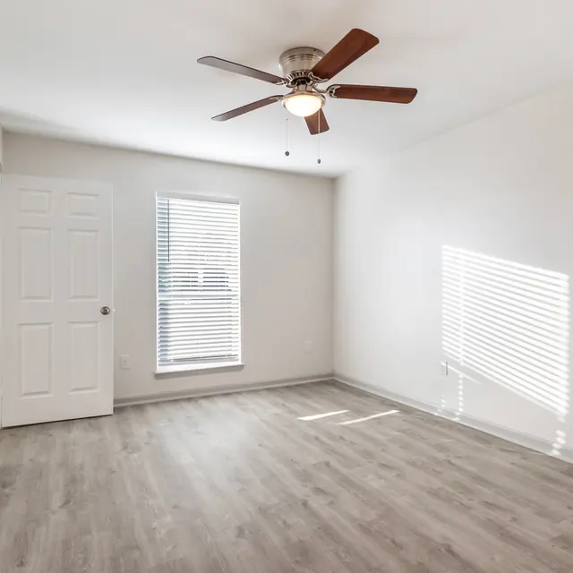 A spacious empty room with light-colored walls and wood-like flooring. There is a ceiling fan with wooden blades and a closed door on one side of the room. A window with blinds allows sunlight to stream in, casting shadows on the floor.