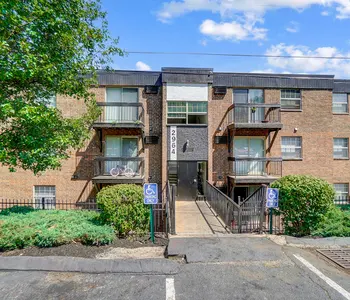 Exterior view of a multi-story brick apartment building with balconies, greenery, and a paved path.