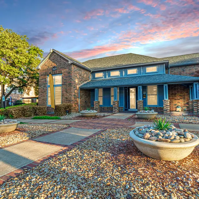 A brick office building with green shutters and a landscaped front yard featuring stone planters and well-kept greenery under a colorful sky.