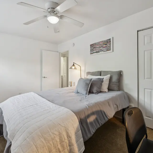 Cozy Bedroom Interior A cozy bedroom featuring a bed with gray bedding, a mirrored dresser, a desk, and a white ceiling fan. The room has a neutral color palette and a piece of wall art.
