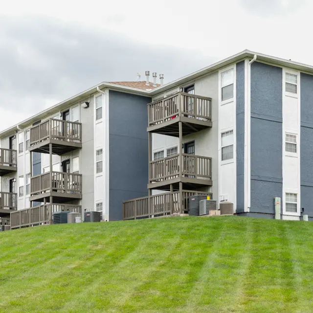 A multi-story apartment building with a blue and white exterior, featuring balconies and a grassy hillside in the foreground.