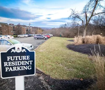 A sign indicating 'Future Resident Parking' in a grassy area next to a parking lot with several parked cars. Trees and a cloudy sky are visible in the background.