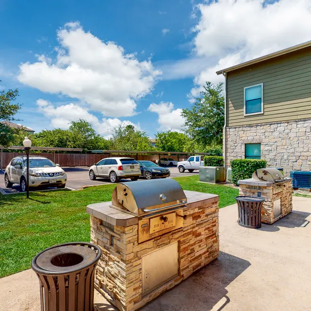 Outdoor Grilling Area Outdoor grilling area with stone grills and parked cars, surrounded by green grass and trees under a partly cloudy sky.