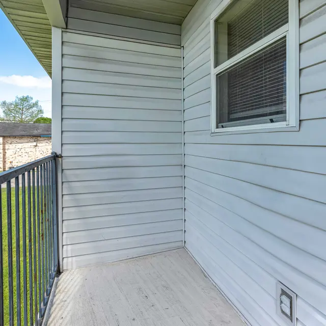 A small balcony with a metal railing, featuring a view of grassy land and nearby buildings. The wall is made of light-colored siding, and there is a window on the right side of the image.