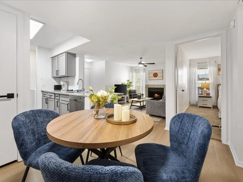 A modern open-concept living space featuring a round wooden dining table surrounded by blue upholstered chairs. In the background, the kitchen is visible with gray cabinets, and a cozy living room with a fireplace is seen through an open doorway.