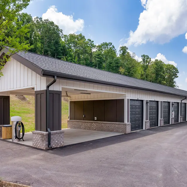A modern storage unit building featuring multiple garage doors, with a paved driveway and greenery in the background.