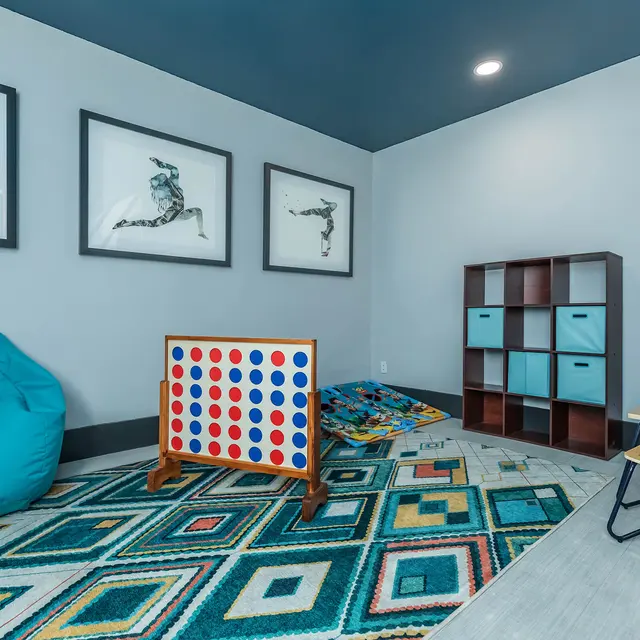 A colorful playroom featuring a large blue bean bag chair, a colorful rug with geometric patterns, a game board, shelves, and framed artwork on the wall.