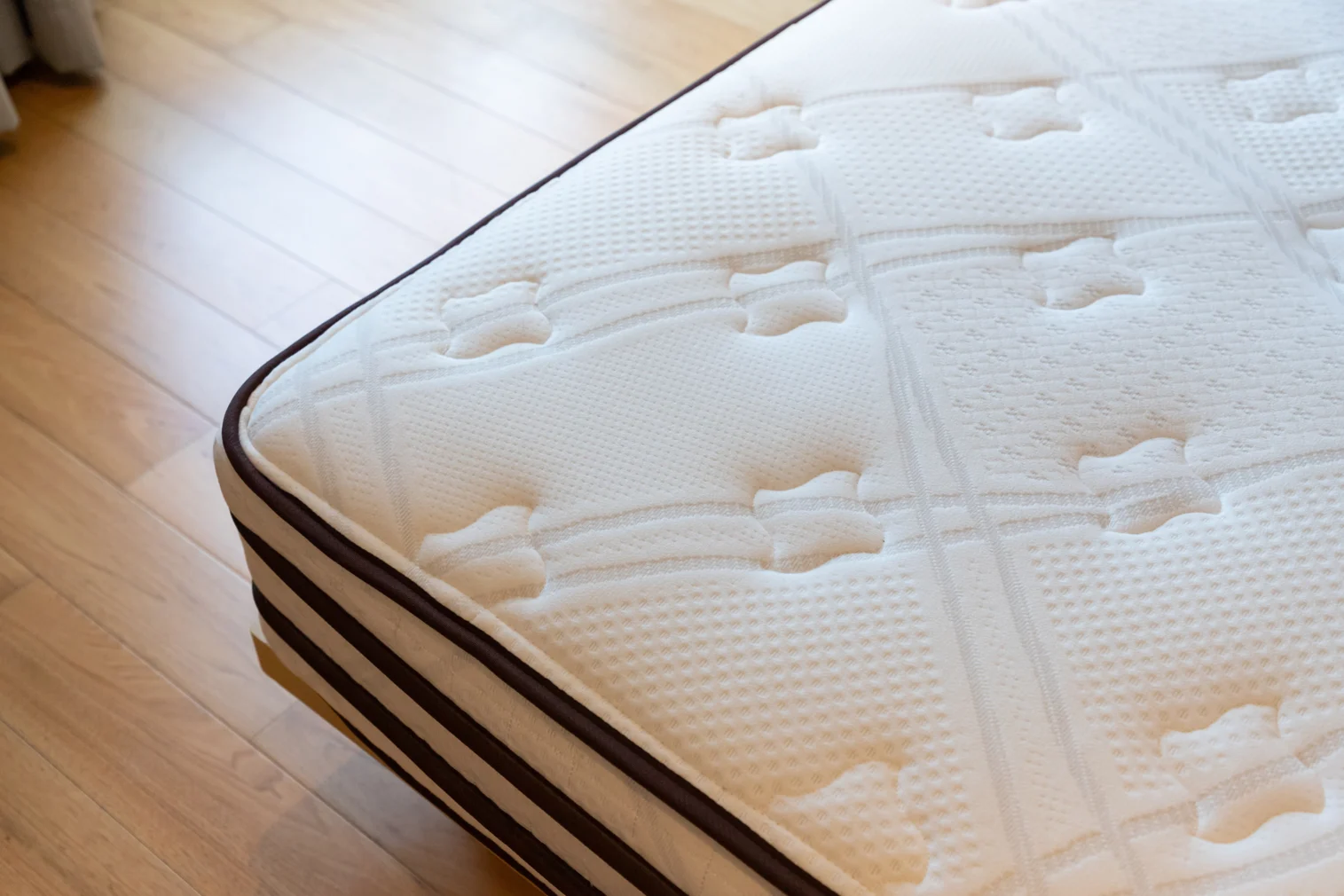 Quilted Mattress on Wooden Floor A close-up view of a mattress resting on a wooden floor. The mattress has a quilted texture with a light-colored fabric surface and dark stripes along the sides.
