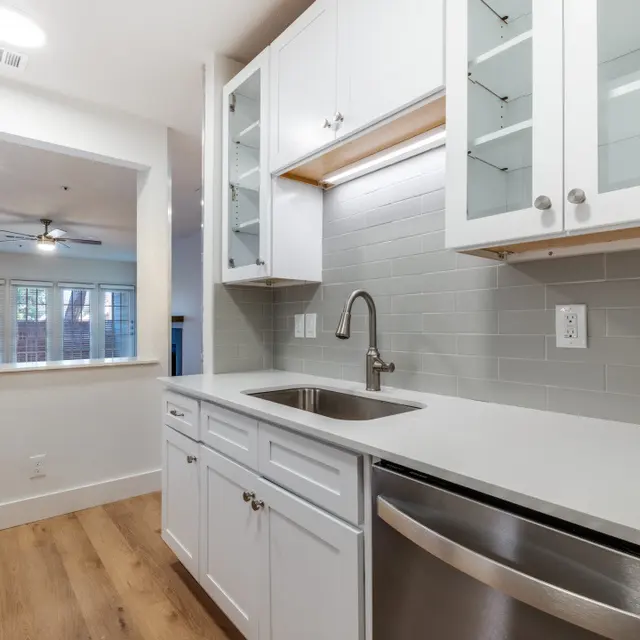 A modern kitchen space featuring white cabinetry, stainless steel appliances, and an undermount sink with a contemporary faucet. The room has light wood flooring and glass-front cabinets.