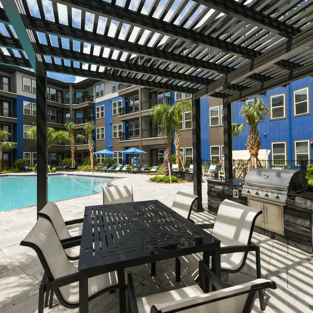 Community Pool and Grilling Area An outdoor community area featuring a pool, grilling station, and seating under a pergola surrounded by palm trees and apartment buildings.