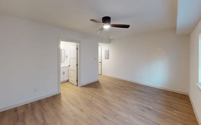 A spacious living room with hardwood flooring, a ceiling fan, and a doorway leading to a bathroom. The walls are painted white and there's a window with blinds.