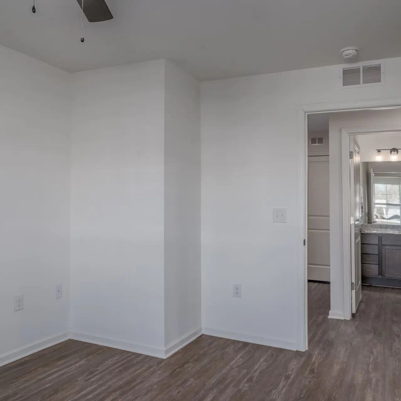 An empty room featuring light-colored walls and a wooden floor, with a doorway leading to a bathroom visible in the background.