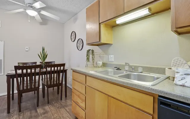 A modern kitchen area featuring wooden cabinets, a double sink, and a dining table with chairs in the background. A ceiling fan is present and the walls are decorated with wall art.