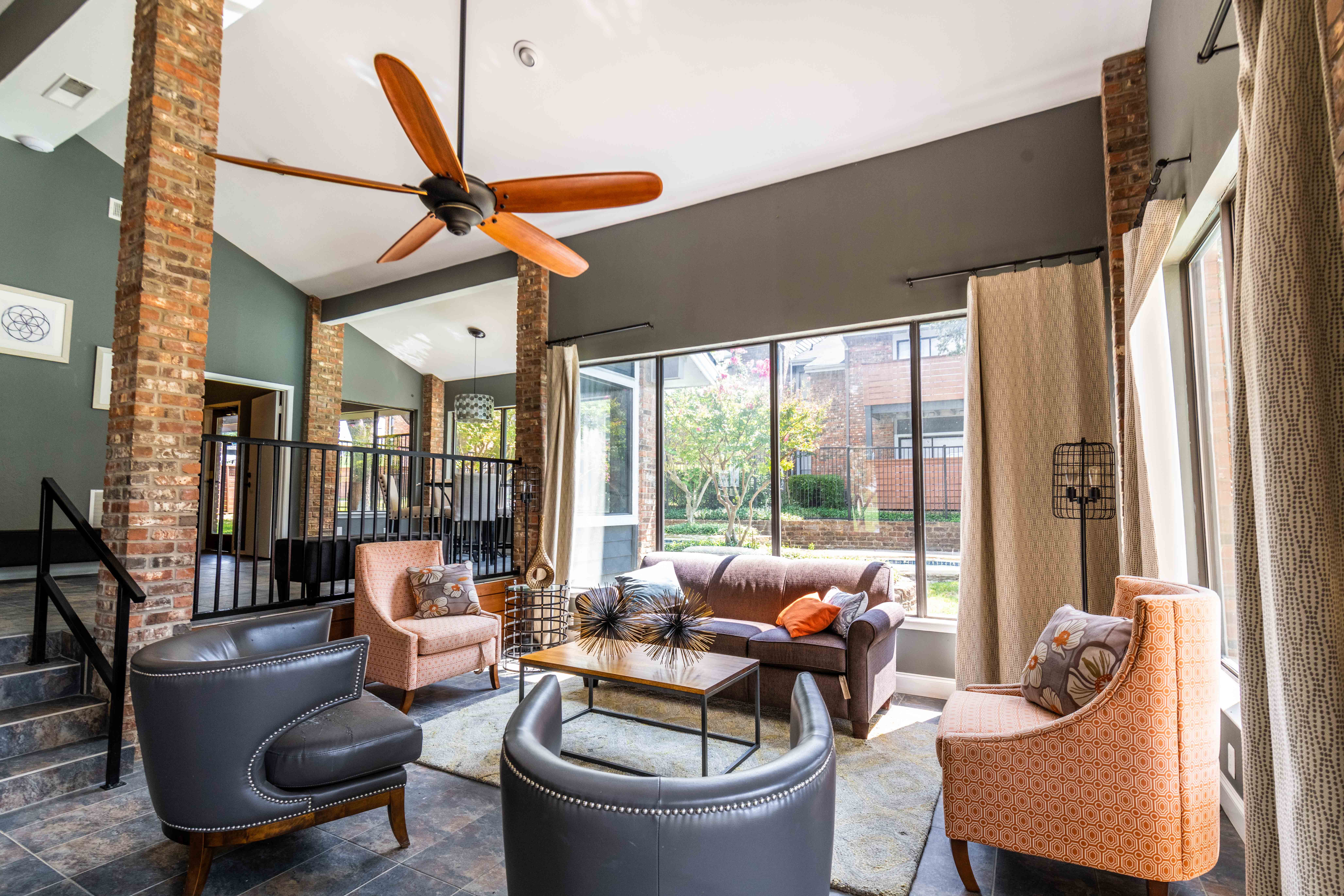 Contemporary Living Room Design A modern living room with large windows, featuring a brown sofa, two armchairs, a coffee table, and a ceiling fan. The walls are a mix of gray and exposed brick, enhancing the contemporary aesthetic.