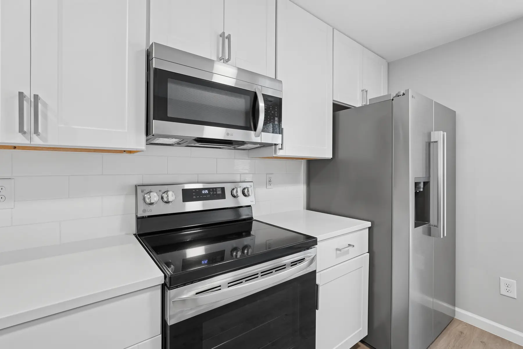 A modern kitchen featuring white cabinets, a stainless steel stove, microwave, and refrigerator. The countertops are light-colored and the walls are painted a soft gray.