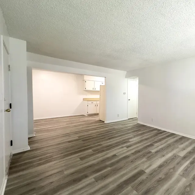 An empty room with new flooring and white walls, featuring a small kitchenette in the background.