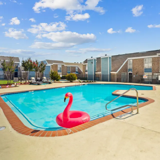 A bright blue swimming pool surrounded by a tan deck, featuring a pink flamingo float. In the background, there are buildings with a modern design.