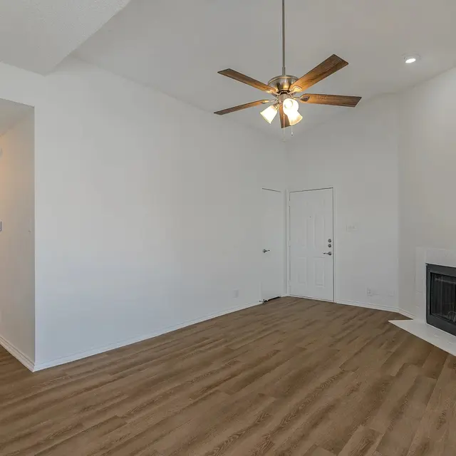 A spacious interior of a living room featuring a ceiling fan, hardwood flooring, and a fireplace. The walls are painted white, and there are doors leading to other rooms.