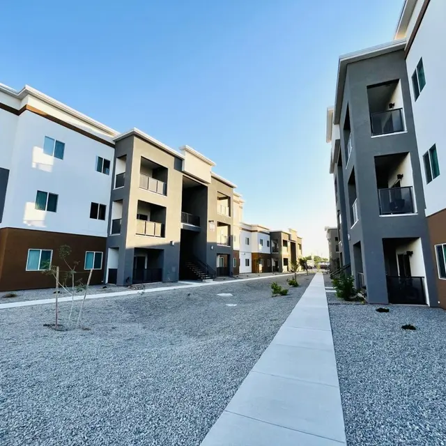 A newly built apartment complex with two rows of three-story buildings on either side of a gravel pathway. The buildings have a modern design featuring a mix of white, gray, and brown exteriors, and the sky above is clear and blue.