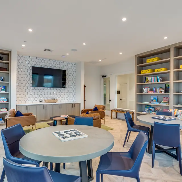 A modern community room with round tables and purple chairs, featuring a television on a geometric wall, shelves filled with books and games, and comfortable seating areas.