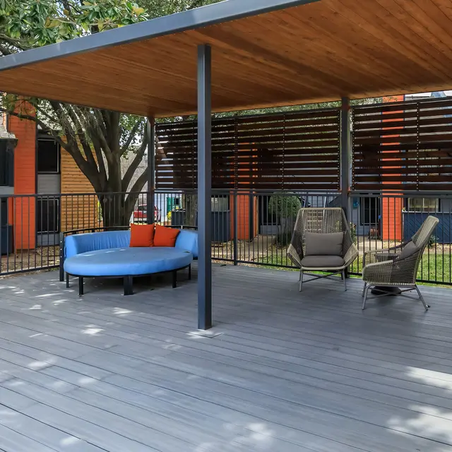 A modern patio area with a blue circular seating arrangement and two lounge chairs, shaded by a wooden roof and surrounded by greenery.