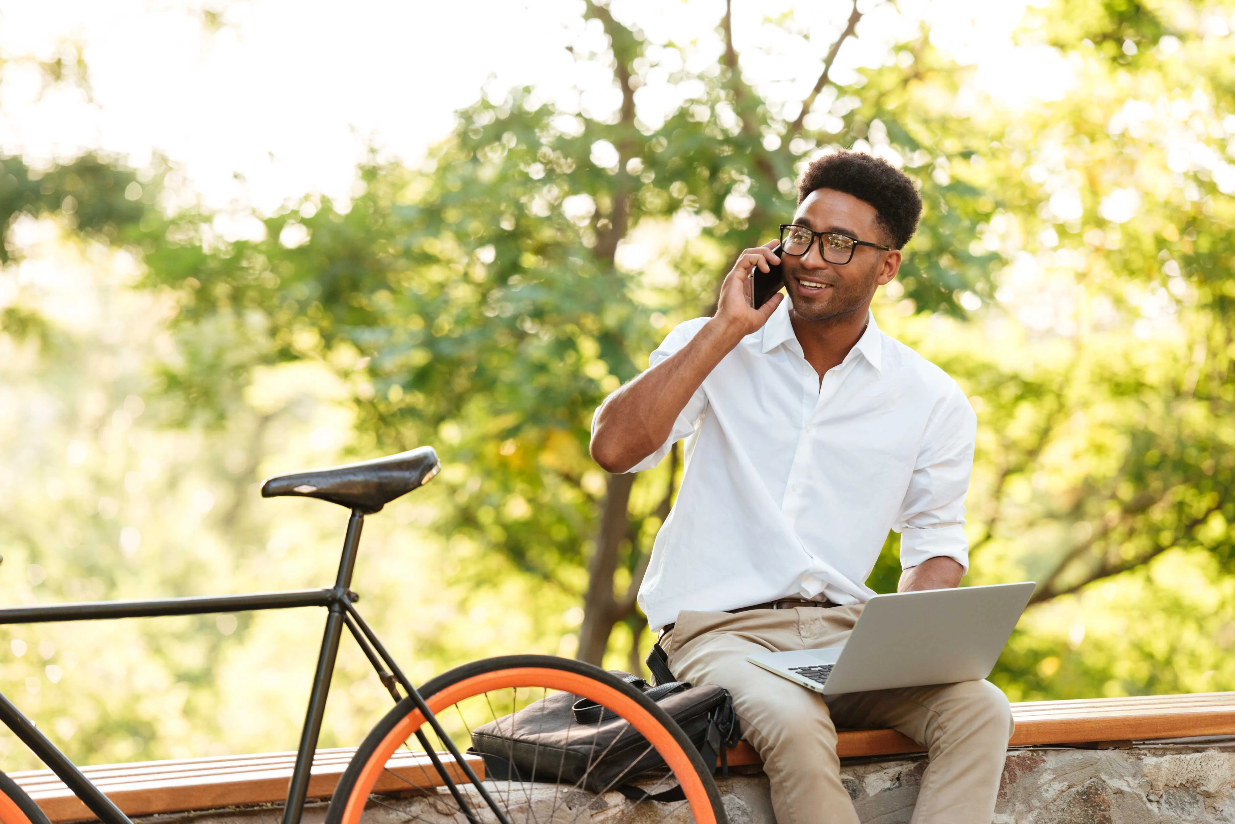 Outdoor Work and Leisure Balance A man in a white shirt and glasses sitting on a wooden bench, holding a phone to his ear, with a laptop on his lap and a bicycle beside him. He is surrounded by greenery, indicating an outdoor setting.