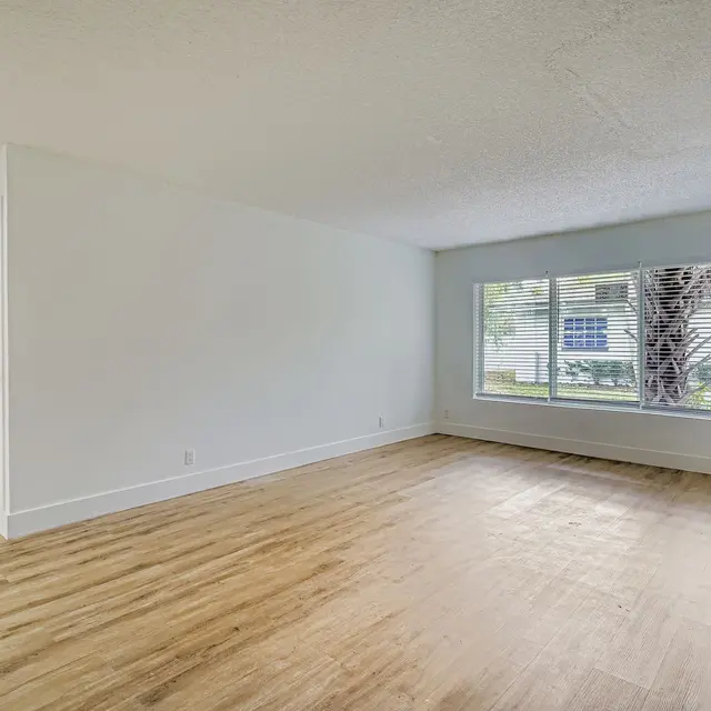 An empty living room with light wood flooring and a large window showing a view of the outside.