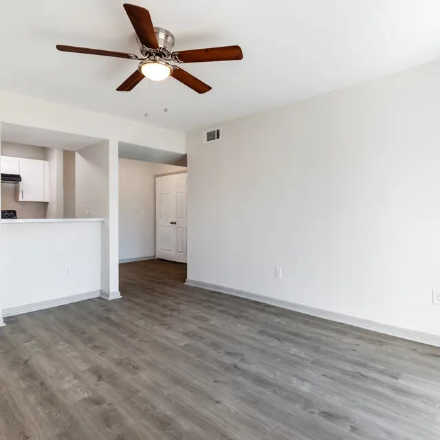 An empty living room with wooden flooring, a ceiling fan, and an open layout connecting to a kitchen area and hallway.
