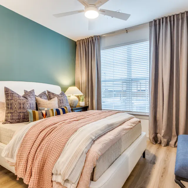 A modern bedroom featuring a white bed with colorful pillows and a warm throw, beige curtains, and a wooden nightstand with a lamp.