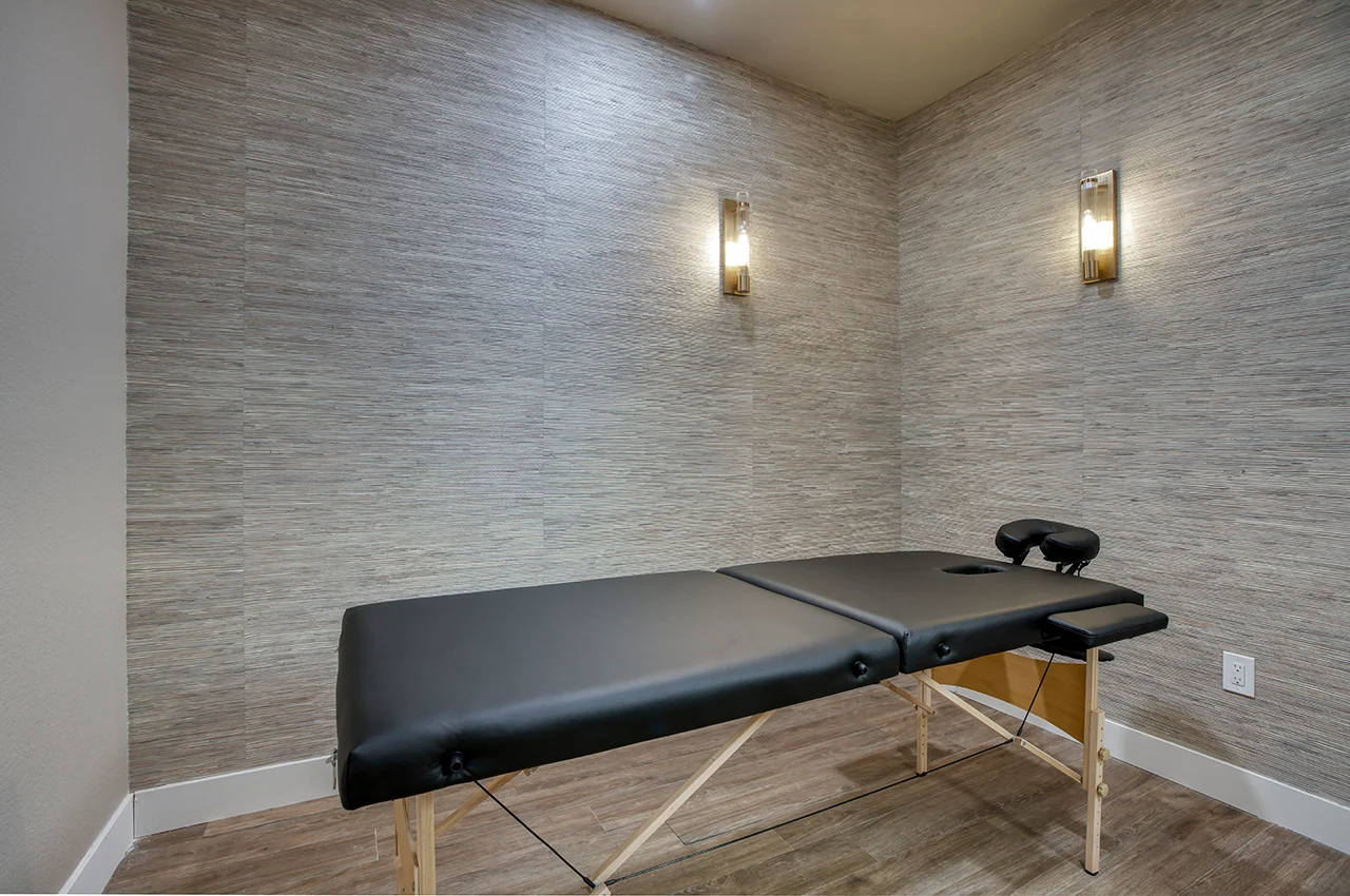 A massage therapy room featuring a black massage table against a textured gray wall, with soft lighting.