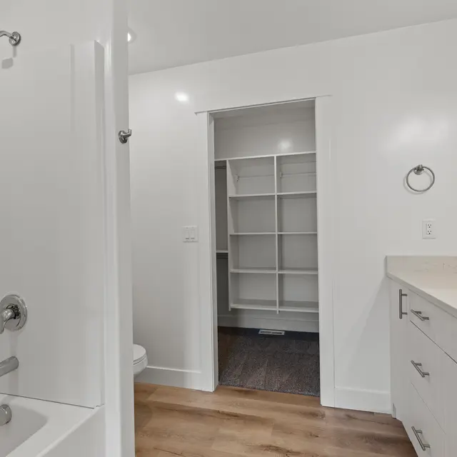 A modern bathroom featuring a shower, sink, and an open storage closet. The walls are painted white, and the flooring is wooden.