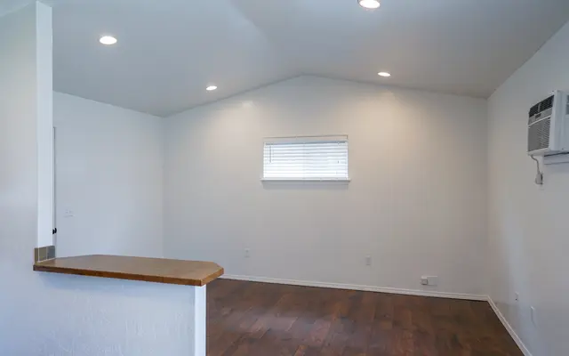 Empty Room Interior An empty interior room with white walls, wooden flooring, and natural light coming through a window. A small counter is visible to the left, and there is an air conditioning unit mounted on the wall.