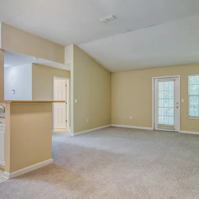 A spacious living area connected to a kitchenette, featuring beige walls, carpeted floor, and large windows.