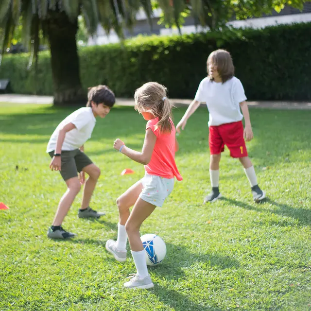 Three children playing soccer on a grassy field, with cones set up for practice.