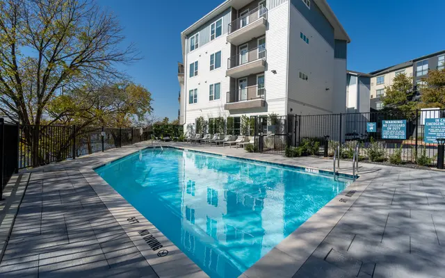 A clear blue swimming pool surrounded by a stone patio and a green tree, with a three-story apartment building in the background.