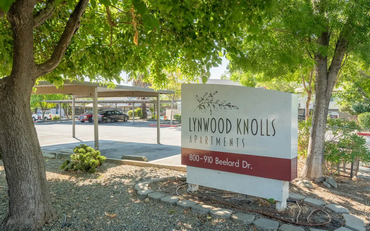 Sign for Lynwood Knolls Apartments surrounded by greenery with shaded parking area in the background.