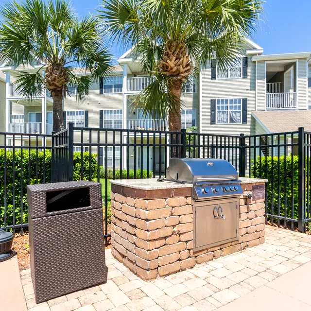 An outdoor grilling area with a stone grill and a trash can, surrounded by lush greenery and an apartment building in the background.