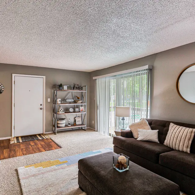 A cozy living room featuring a dark brown sectional sofa with decorative pillows, a round mirror on the wall, wooden floors, a shelving unit with decorative items, and sliding glass doors leading outside.