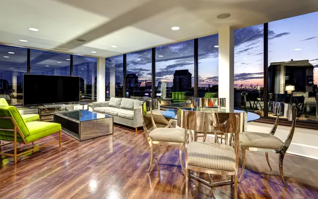 A modern living room with large windows showing a sunset view. The room features a green armchair, a gray sofa, a glass coffee table, and a dining table with metallic chairs.