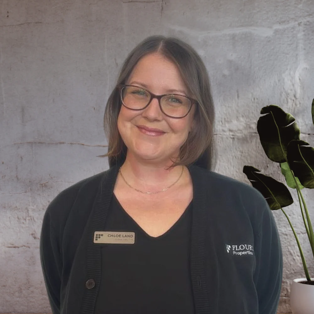 A smiling woman with glasses stands in front of a textured background, wearing a black blazer with a nameplate.